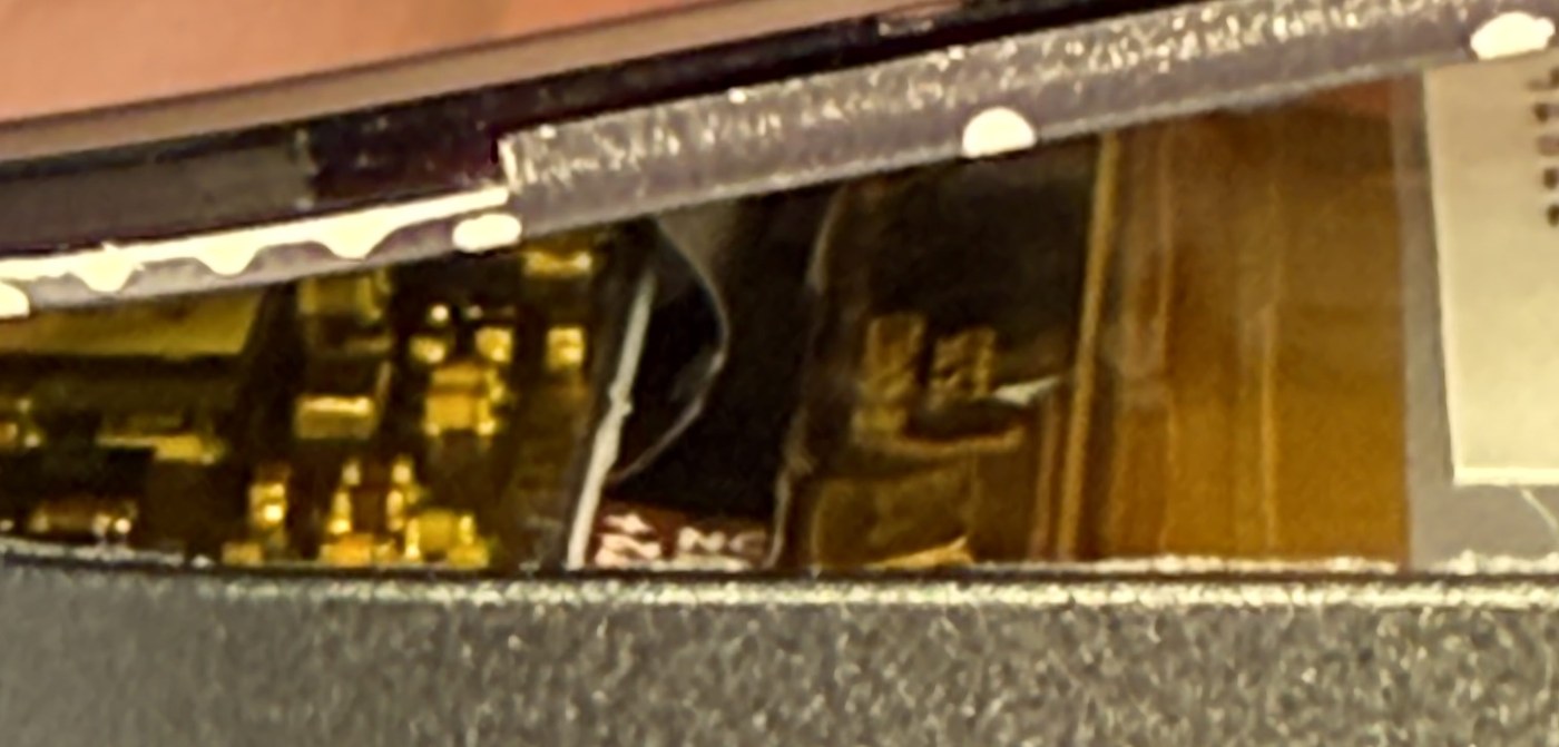 Close-up under the lifted Trezor Safe 7 display edge showing the adhesive line, the gold mainboard components and the battery beneath