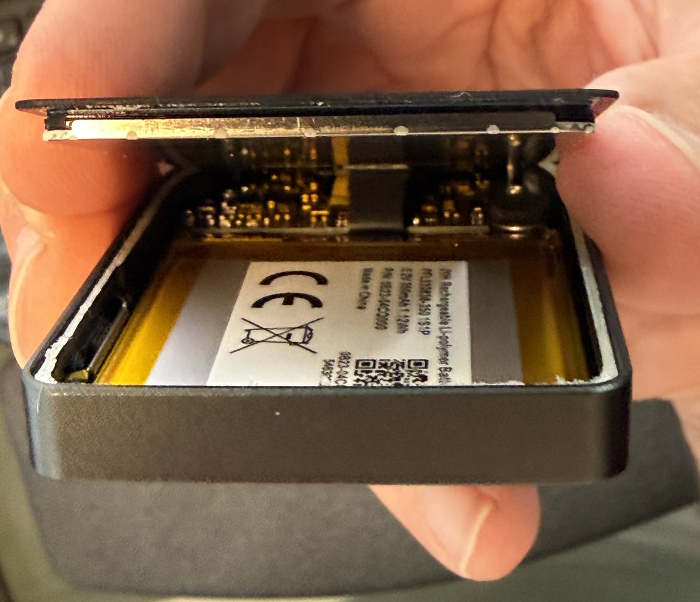 Hand holding the opened Trezor Safe 7 with the display hinged up from the frame, exposing the gold mainboard and the battery label beneath it