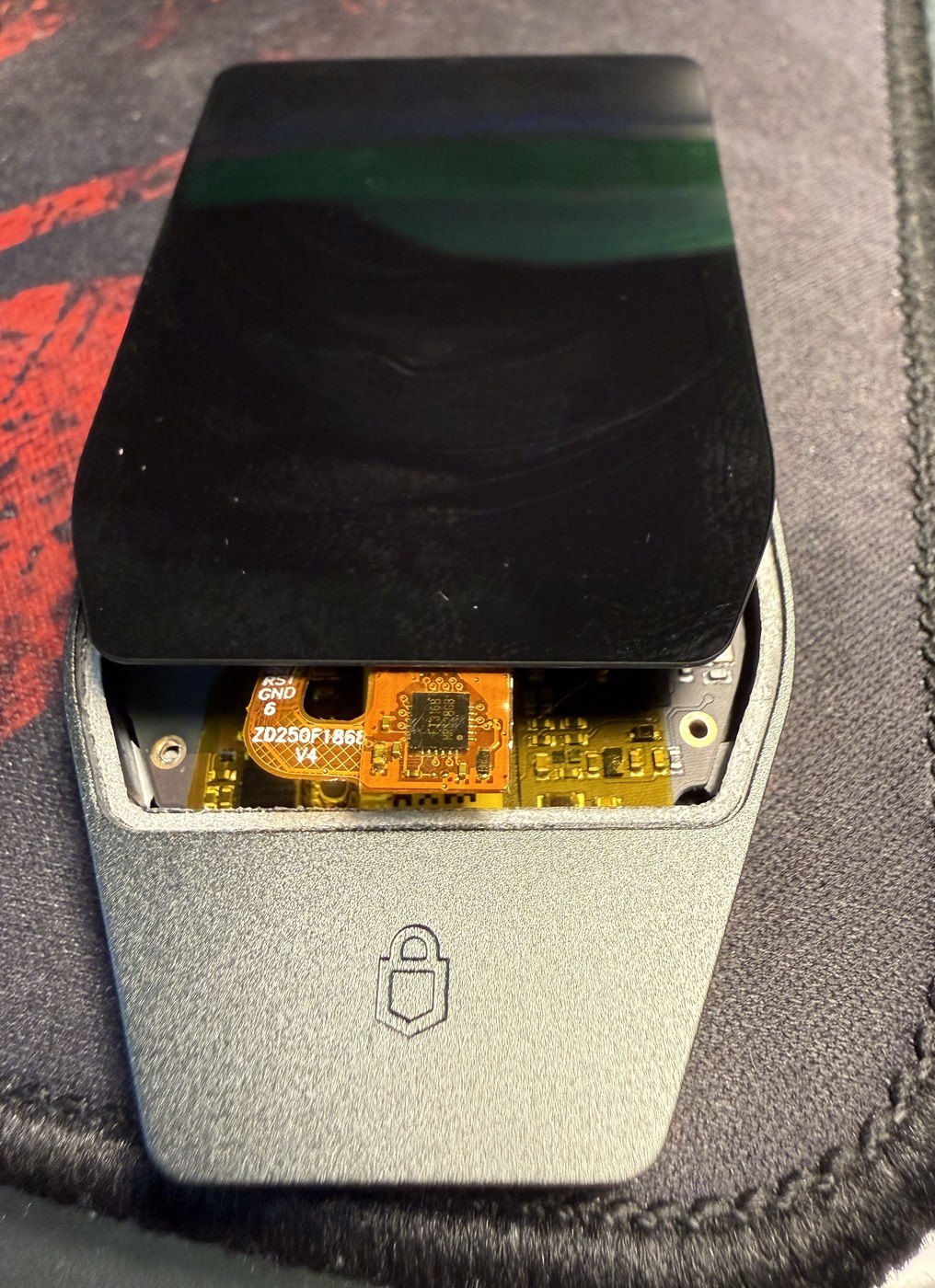 Trezor Safe 7 display lifted clear of the lower frame, exposing the orange display flex cable and its driver chip above the mainboard