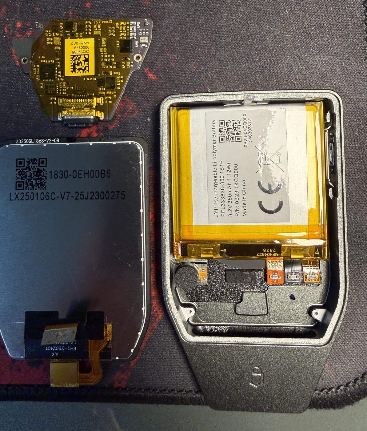 Trezor Safe 7 disassembled: the TS7 rev.D mainboard at top left, the display panel with its flex cable below it, and the open aluminium enclosure with the lithium polymer battery at right
